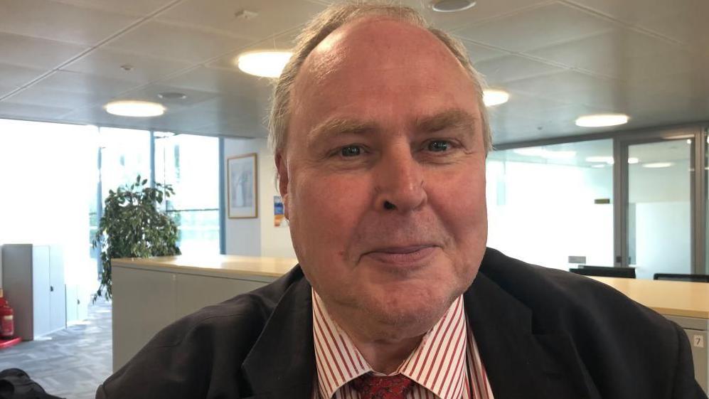 A man is in an office smiling at the camera and is wearing a suit and striped red and white shirt, as well as a red patterned tie.