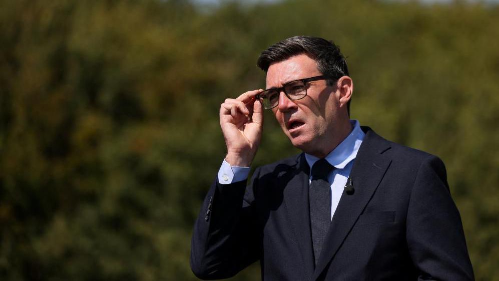 Prime Minister Andy Burnham talking while adjusting his glasses