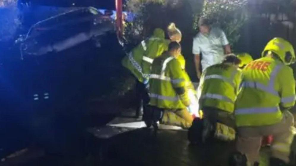 A silver VW Passat poking through a hedge it has crashed through.  In the foreground are firefighters