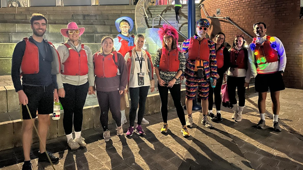 About ten canoers standing on the wharfside at night, lit up by lamplight wearing lifejackets and some are in bright clothing such as a pink wig, pink brimmed hat and fairy lights
