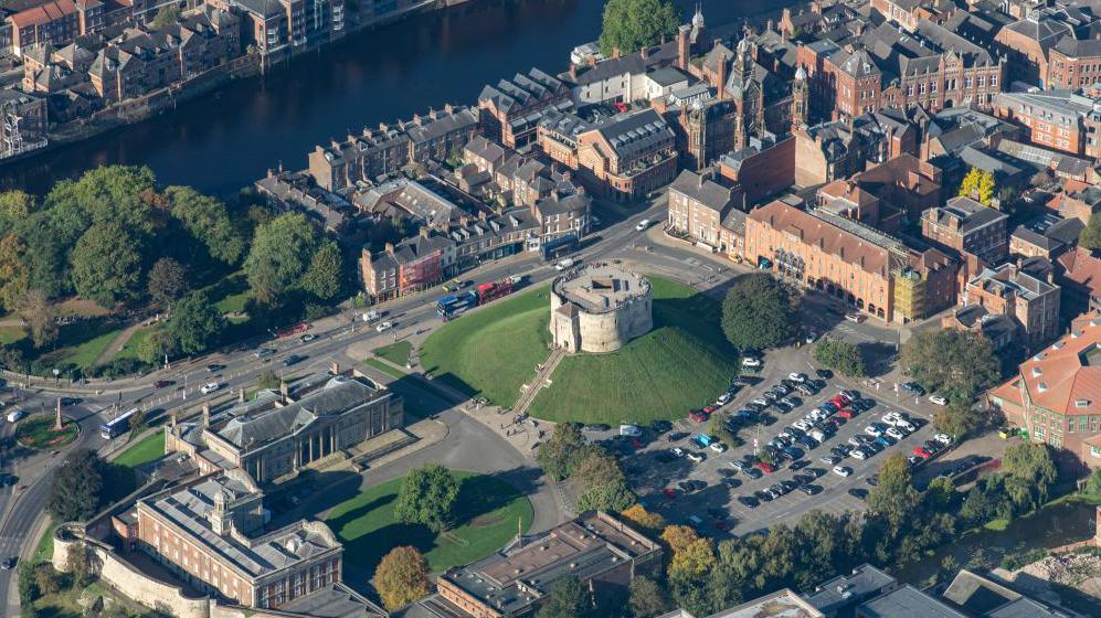 An aerial view of the Eye of York with Clifford's Tower, a medieval tower sitting on a mound, in the centre of the image, with a car park to the right of the image behind the tower. To the left of the image is a group of Georgian buildings including the city's Crown Court, and the Castle Museum