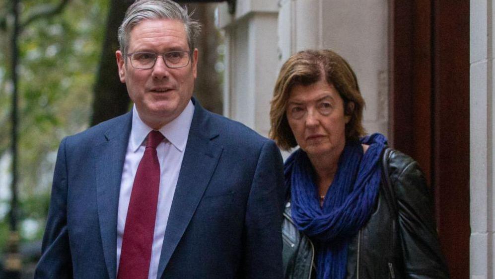 Keir Starmer and Sue Gray. Starmer is wearing a grey suit jacket and a white shirt with a red tie. Sue is wearing a leather jacket and a blue scarf.