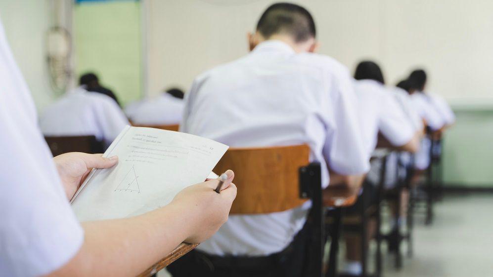 Generic image of uniformed students sitting in an exam hall - a student is holding up the exam paper which has a graph question on it