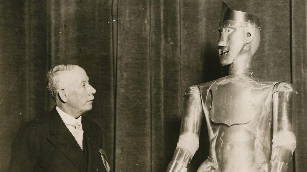 A black and white image of a man in a suit stood next to a metal robot. The robot has a face with eyes, ears, nose, and a mouth.