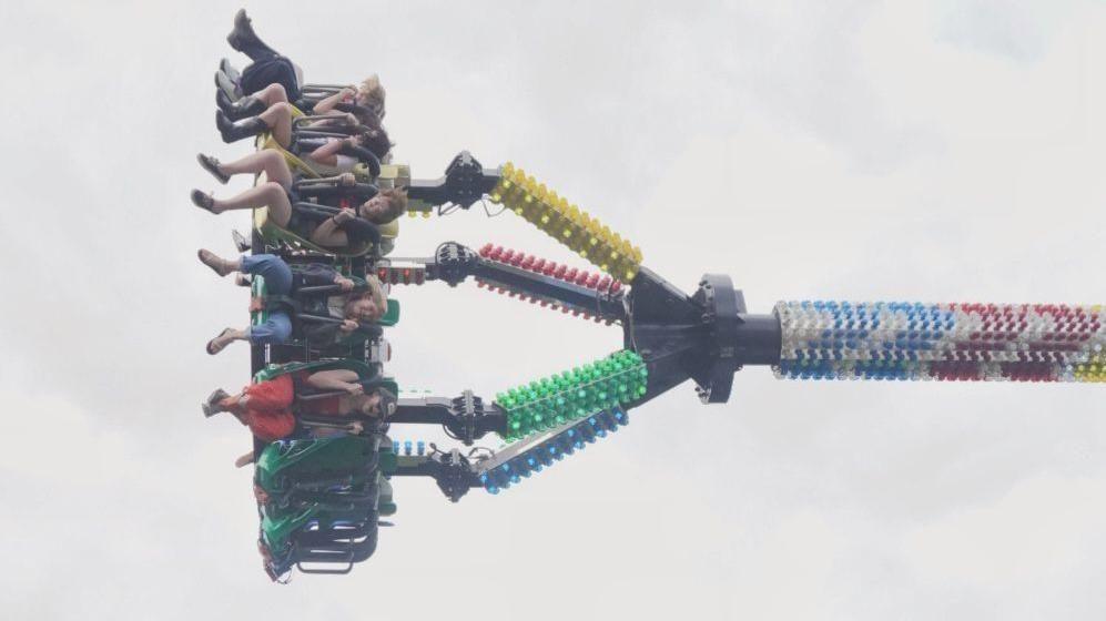 People in mid-air strapped into a fairground ride which has carried them near-horizontal into the air with colourful lights along its structure.
