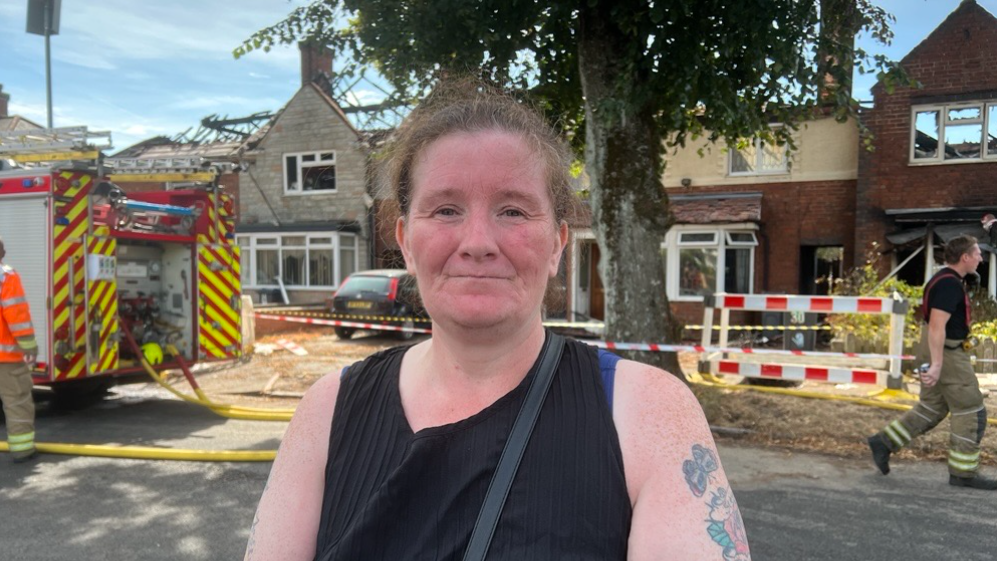 A woman with brown hair tied back with tattoos on her left arm. She is wearing a black tank top and is standing in front of a fire engine which is outside a house damaged by fire with no roof