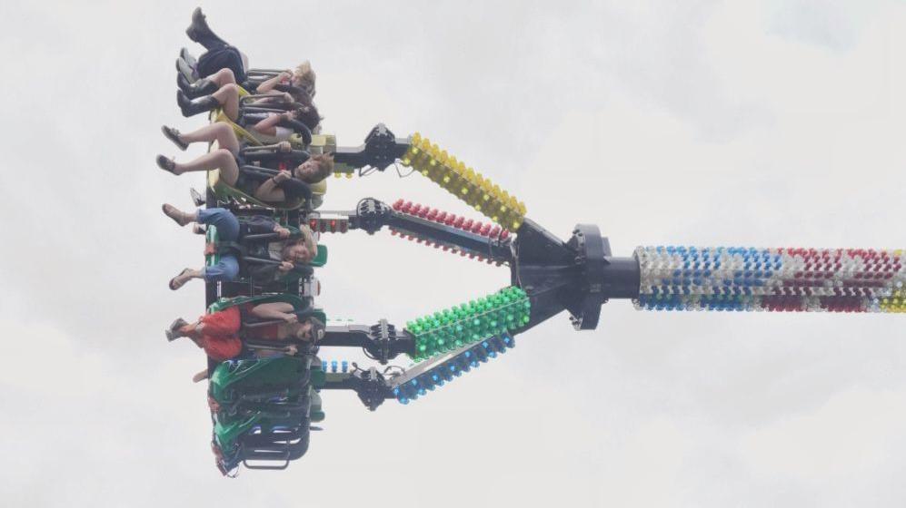 People in mid-air strapped into a fairground ride which has carried them near-horizontal into the air with colourful lights along its structure.