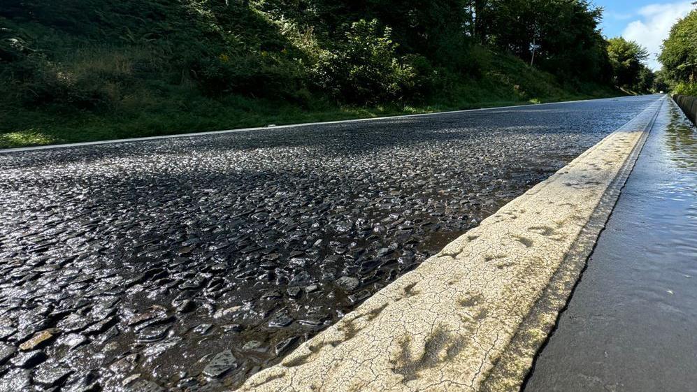 A dirty looking white line along the side of a road, which is wet. There are green embankments at wither side of the road and blue sky up ahead.