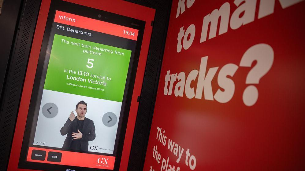 A red screen with passenger information on and a man doing British Sign Language