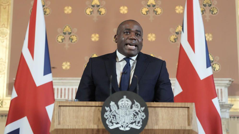 UK Foreign Secretary David Lammy in the foreground dressed in dark blue suit with the national flag of the UK in the background in a room where he gave a speech