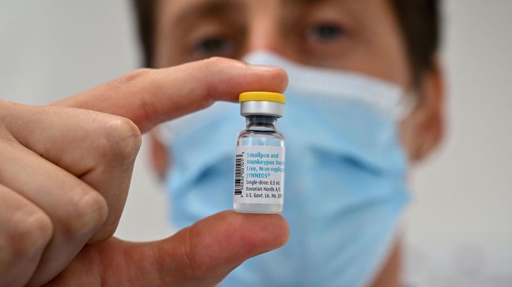 A scientist wearing a blue surgical mask holds a small glass vial of mpox vaccine. 