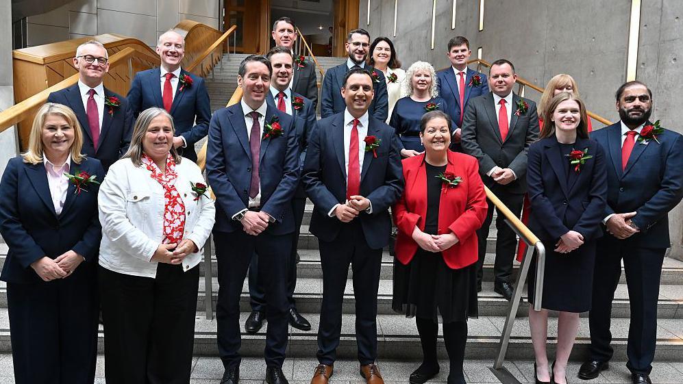 Group of around 15 people wearing business attire and red floral lapel pins pose for a photograph on a staircase inside a modern parliamentary-style building. The group is arranged across several steps, with handrails, concrete walls and wooden architectural features visible in the background.
