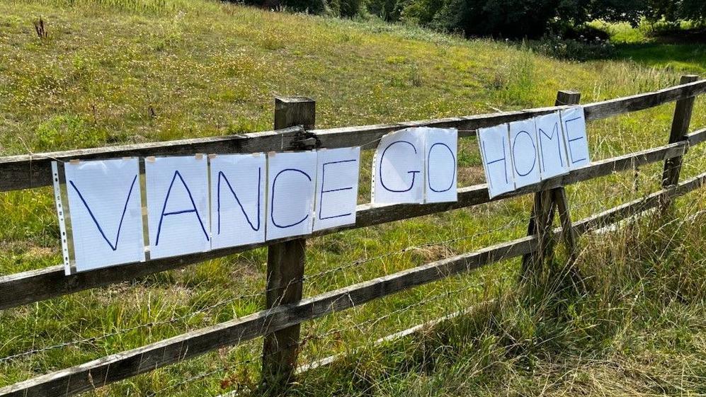 The words "Vance go home" stapled on a fence. Each letter is written on a white sheet of paper. It's a sunny day.