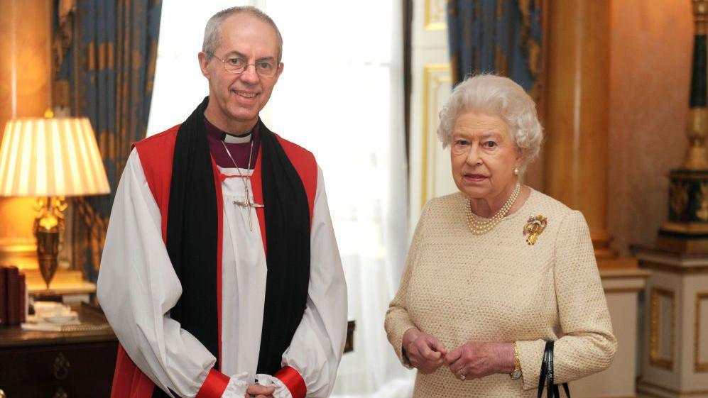 Archbishop Justin Welby, in red, white and black robes, stands in a large room beside Queen Elizabeth II, who is wearing a beige dress and pearls. 