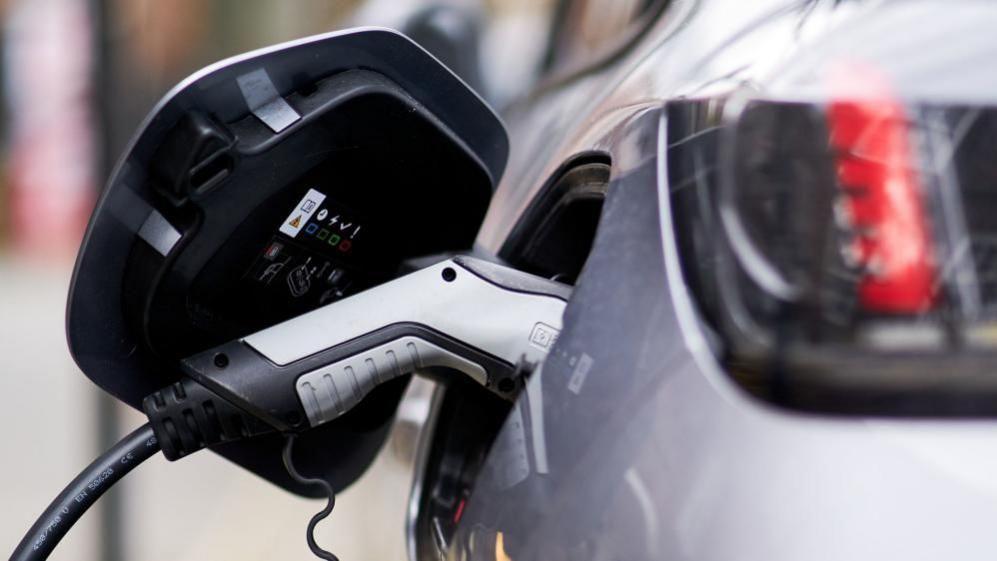 An electric vehicle charge point. A silver car with a silver handled charger.