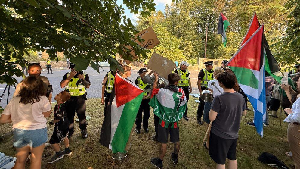 A group of protesters, many waving Palestine flags standing behind a row of police officers
