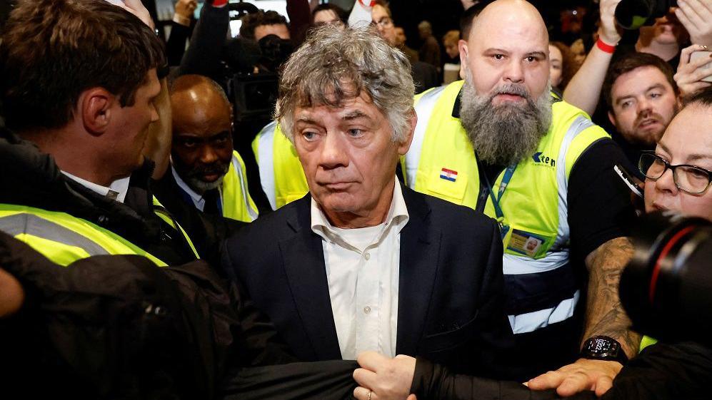 Gerry Hutch has salt and pepper hair and blue eyes and is smiling. He is wearing a dark grey suit jacket and open-necked shirt and is surrounded by photographers