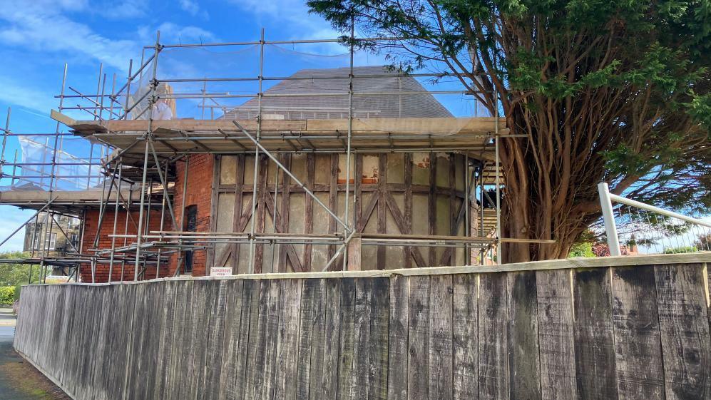 Bramcote Tennis Pavilion in Scarborough surrounded by scaffolding. The 19th Century pavilion is a red-brick building with Tudor style wooden cladding around one section. A wooden slatted fence stands in front of it, and a large Conifer tree is to the left.