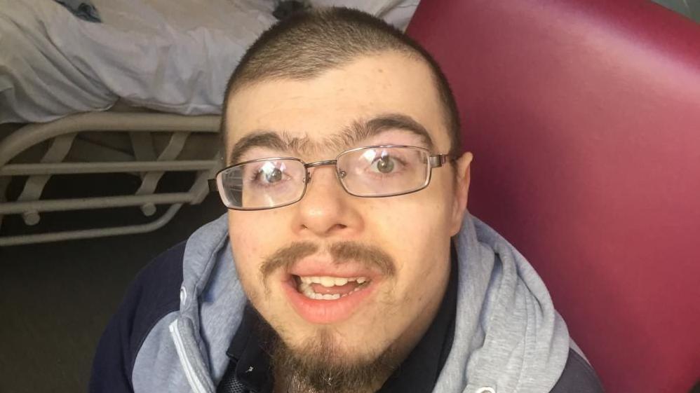 Jordan Tooke, wearing a black and grey hoodie and tee-shirt. He is wearing glasses, is smiling, and sports a goattee beard and moustache, with close cropped hair. He is sitting in a burgundy-coloured vinyl covered arm chair, which is next to a hospital bed.