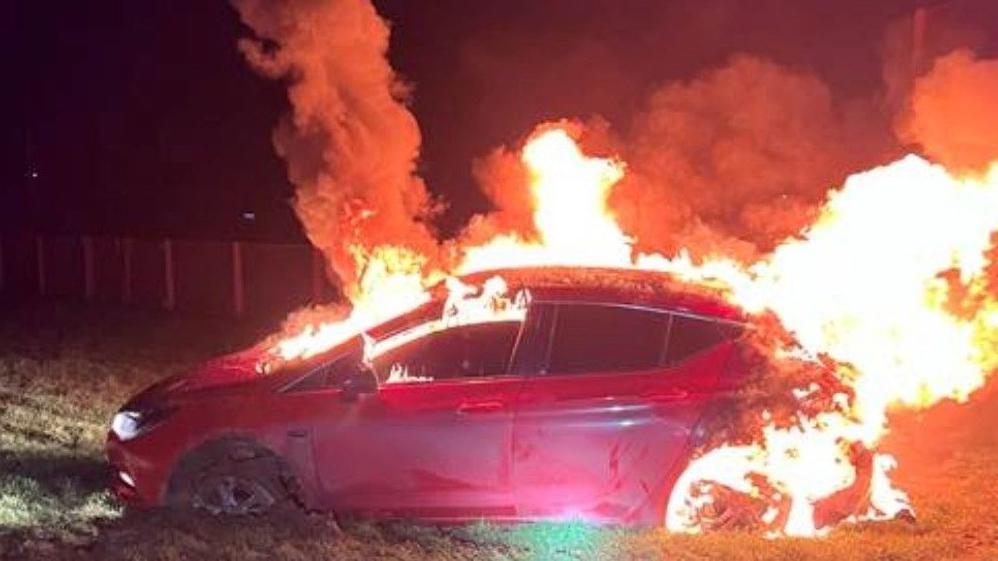 A red car, with flames coming out of the roof, windows and rear. It appears to be in a grassy area, with a set of fence panels visible in the distance. The photo was taken in the dark.