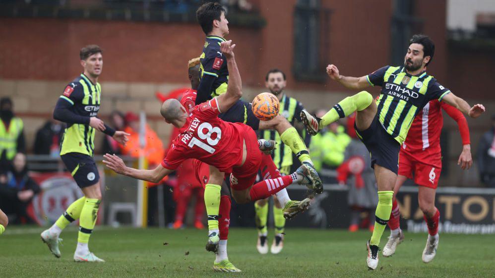Leyton Orient's Darren Pratley and Manchester City's Ilkay Gundogan go in for a tackle, with Orient players wearing all-red kits and City in fluorescent yellow and black striped shirts, black socks and fluorescent yellow socks