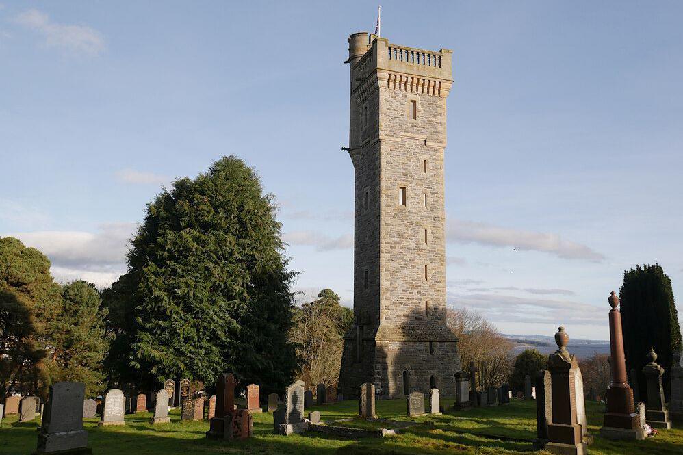 The stone-built tower rises from a graveyard. There is a small turret at the top of the tower and a flag pole. There are trees in the graveyard.