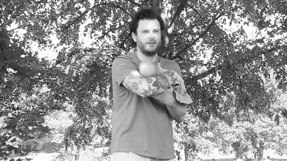 In a black and white image, James can be seen holding newborn daughter Seren in his arms, as he stands under a tree. He wears a short sleeved t-shirt.