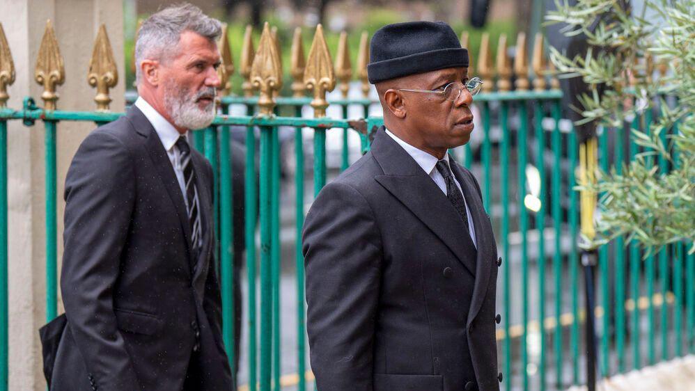 Roy Keane wears a dark suit and has white hair with a beard. Ian Wright wears a black hat, glasses and a dark suit. They are both walking and a green and gold gate is in the background.