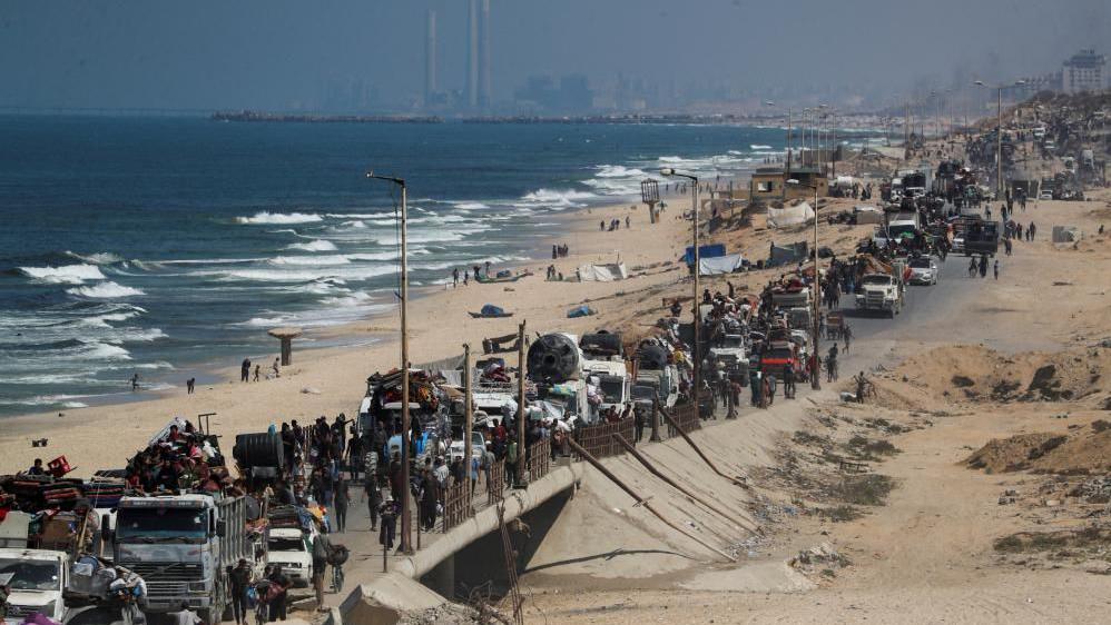 A long line of vehicles and trailers snake along a road, heavily packed with everything they can manage, as people flee south. The road is beachside, and you can see the ocean on the left, and in the distance is Gaza City.