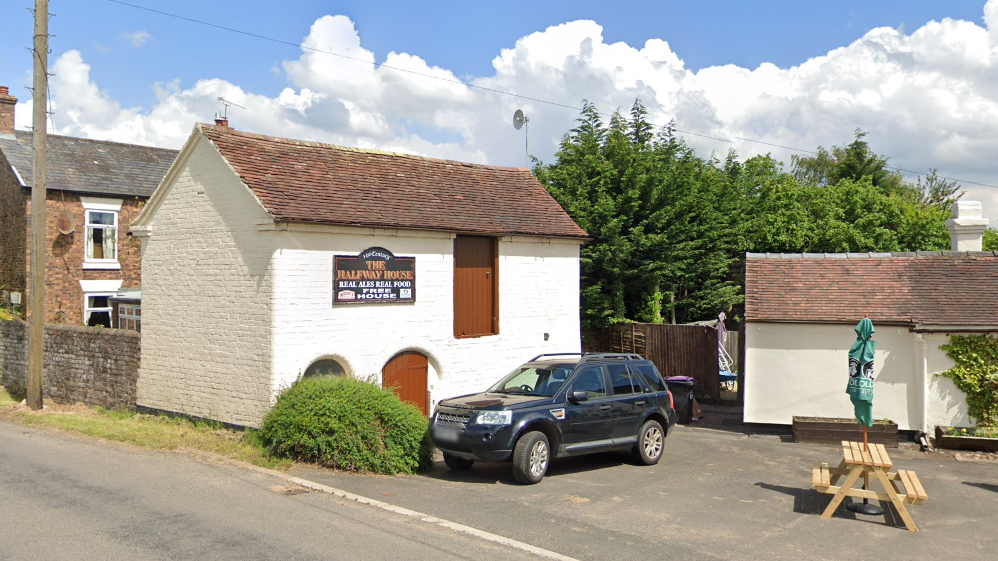 The outside of a white pub building that has brown wooden doors. There is a sign that reads "the halfway house". there is a car parked in front of it and a light wood bench with blue/green umbrella