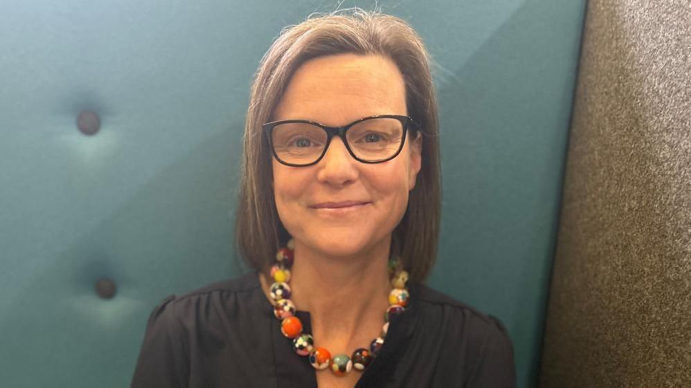 A headshot of a woman who is sitting on a blue sofa. She is wearing a black blouse and a chunky necklace that is made of yellow and orange marbles. She has brown hair that is bobbed and is wearing black glasses.
