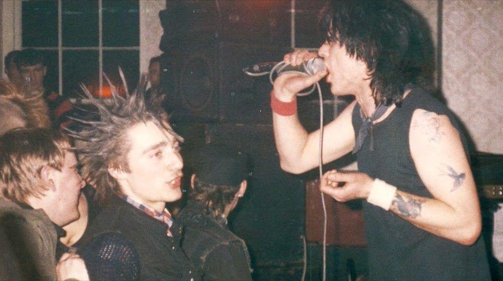 A man with shoulder-length black hair sings into a microphone, with speakers in the background and people in the crowd watching him, including a man with spiky hair