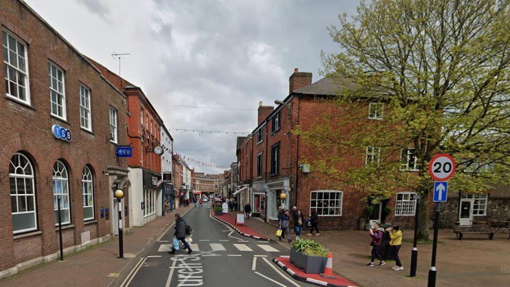 A zebra crossing with a high street stretching into the distance behind it. There are pedestrians walking on a paved area either side and one crossing the road.