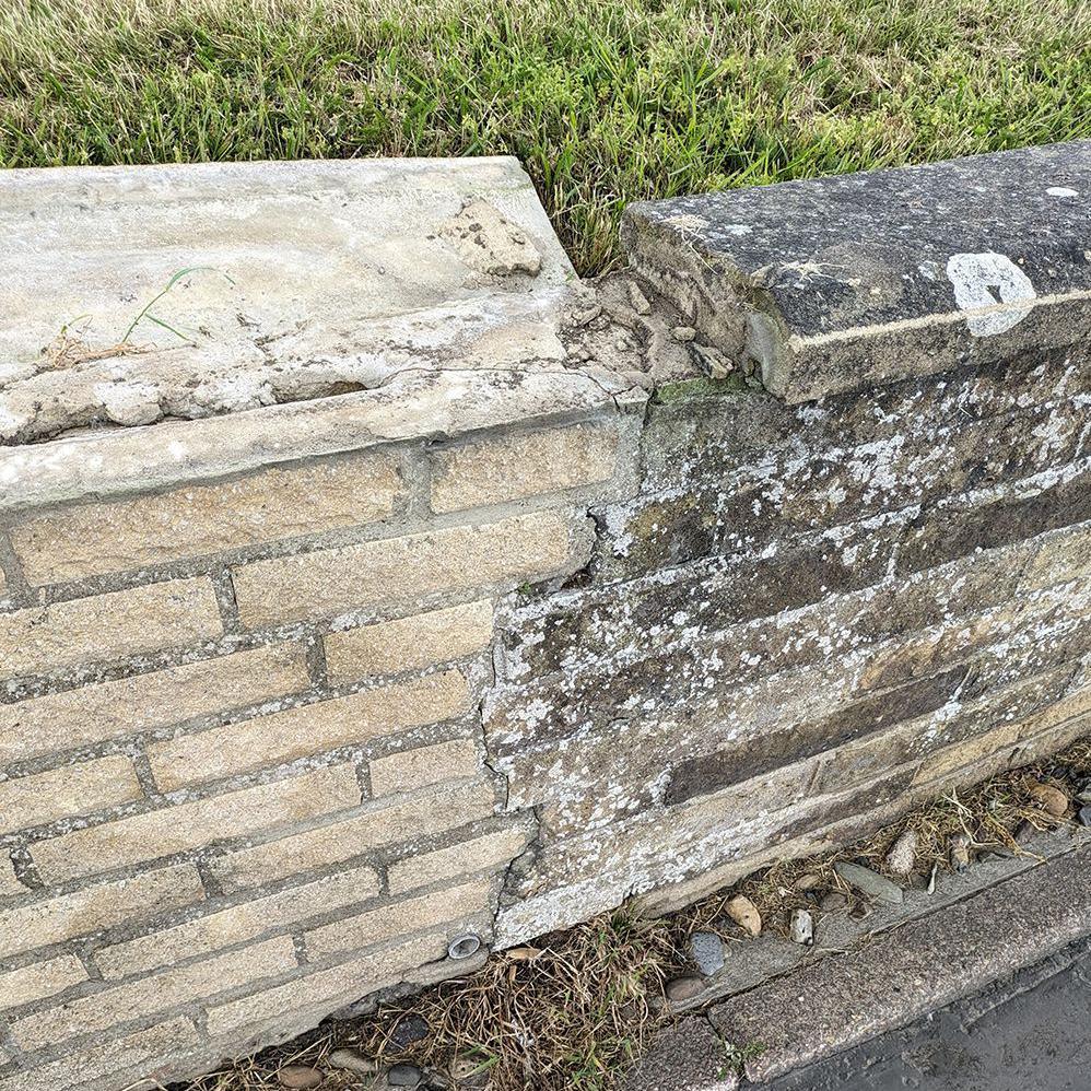 A low wall with a vertical crack in the middle going from top to bottom. The left half of the wall is beige stone while the right shows dark discolouration and mottling on the brickwork. A section of the top is also missing.