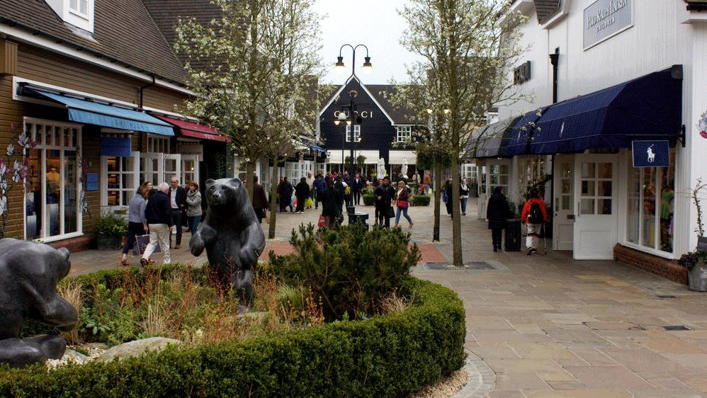 The bear statues at Bicester Village, surrounded by plants and greenery. Shoppers walk the cobbled streets of the outlet village. A Gucci store is in the background.