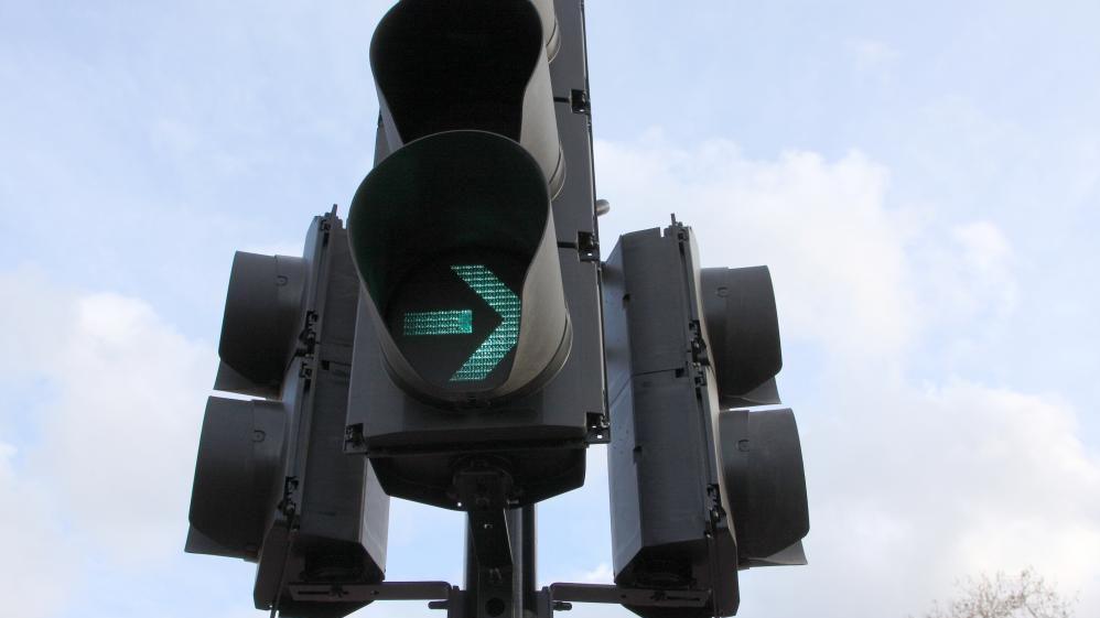 A close up of traffic lights. The signal facing the camera is lit green with an arrow pointing to the right. There is a tree in a background and there are clouds in the sky.