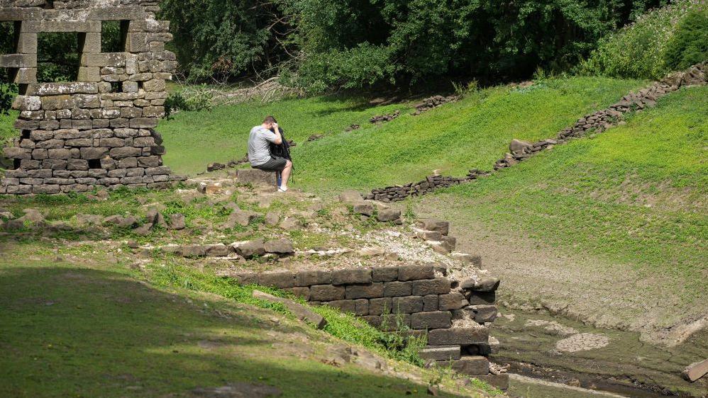 Man in shorts peers into a rucksack as he sits astride a stone next to the ruins of a building. The area has short grass growing around it but is actually normally underwater and has been uncovered by falling reservoir levels.