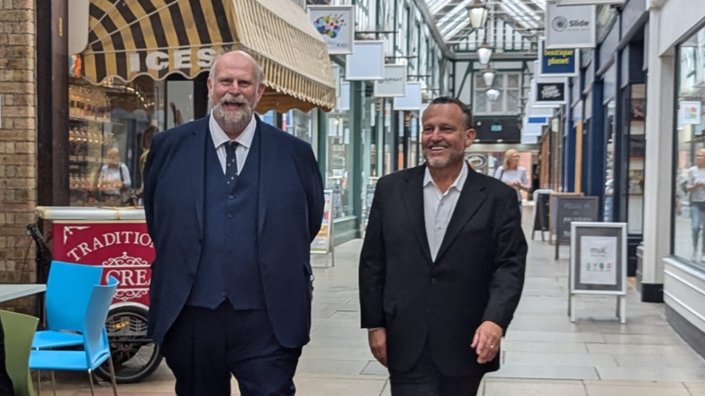 Tom Wootton and John McReynolds walking down Bedford's Arcade shopping centre, wearing dark suits and smiling