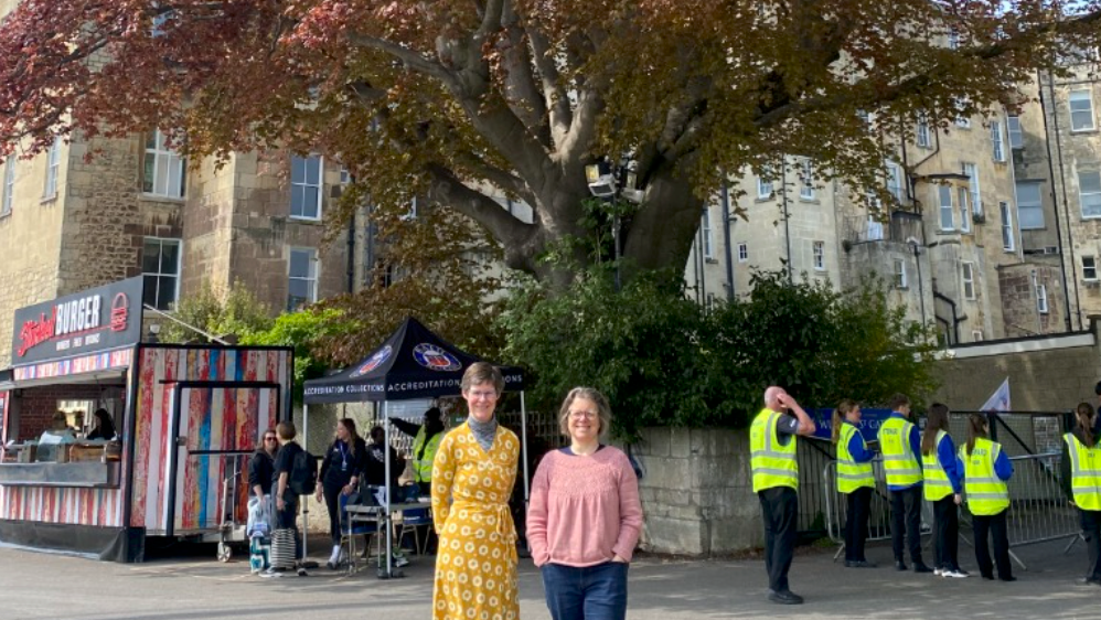 On the left is green councillor Saskia Heijltjes wearing a yellow dress and on the right, Joanna Wright who is wearing a light pink blouse and blue trousers. They are stood in front of the protected copper beech which has a thick trunk and orange-brown leaves.