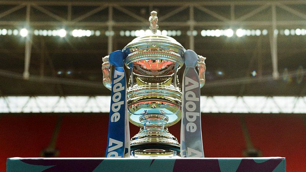 The Women's FA Cup trophy on a plinth with the red seats of Wembley Stadium in the background.