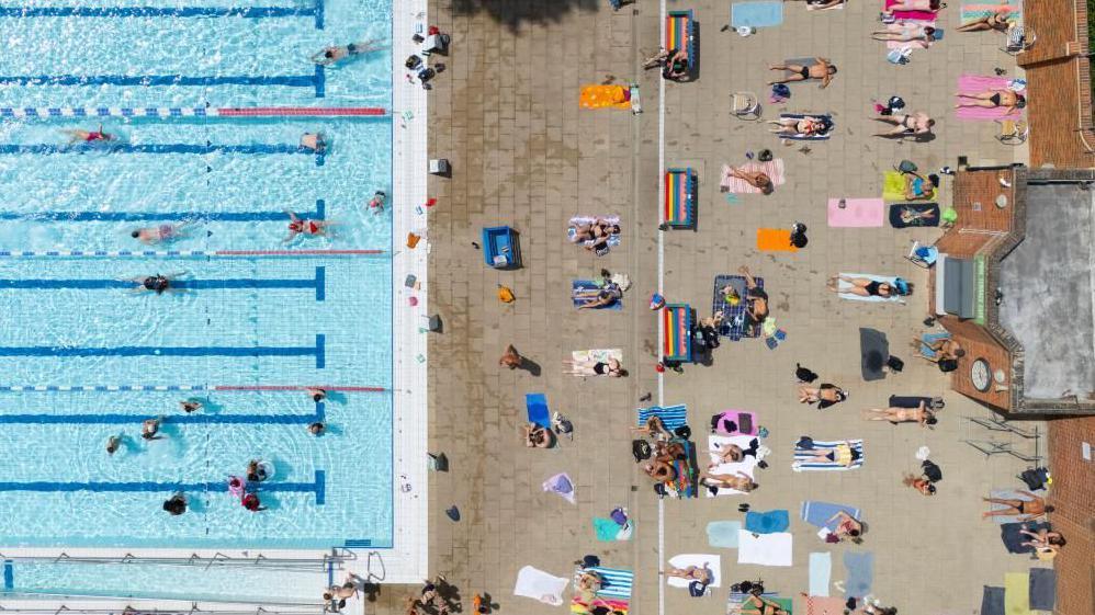 People sunbathe next to an outdoor lido in an aerial shot.