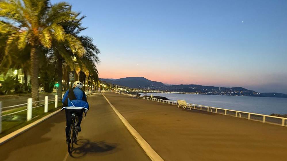 A cyclist wearing a blue jacket cycles along the seafront in Nice, France, at dawn. The sky is blue with a tint of pink and palm trees line the prom.