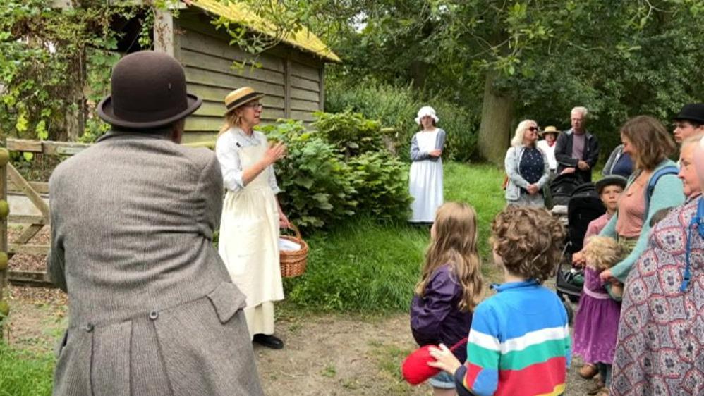 Guides in period costume explain the workings of the farm to a group of adult and child visitors by a wooden gate and building.