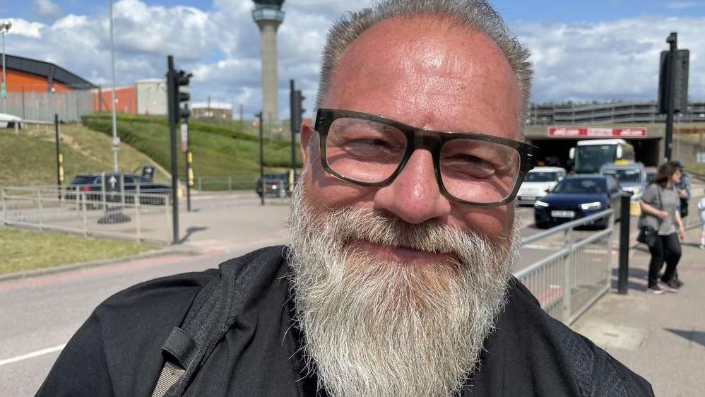 Chris Smith has swept-back hair and a beard. He is wearing glasses, and a black top and rucksack. He is standing outside with a road and air traffic control tower in the background.