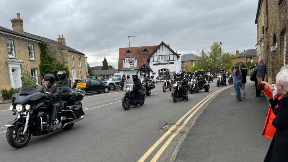 Lots of motorcycles are travelling down a road, and there are people watching from the pavement