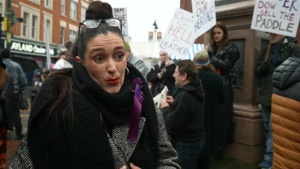 A woman with black hair pulled into a bun looks at the camera as she talks. Behind her are groups of protestors with campaign signs, standing in Walsall town centre. She wears a black scarf, and a black and white striped coat.