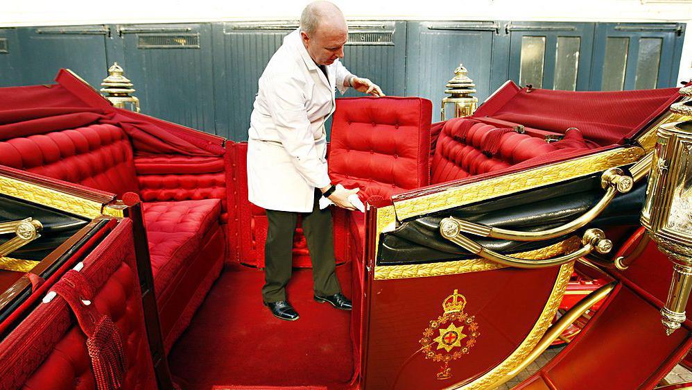 A man in a white coat cleaning the bright red interior of a coach without roof, known as a landau inside the Mews buildings. 