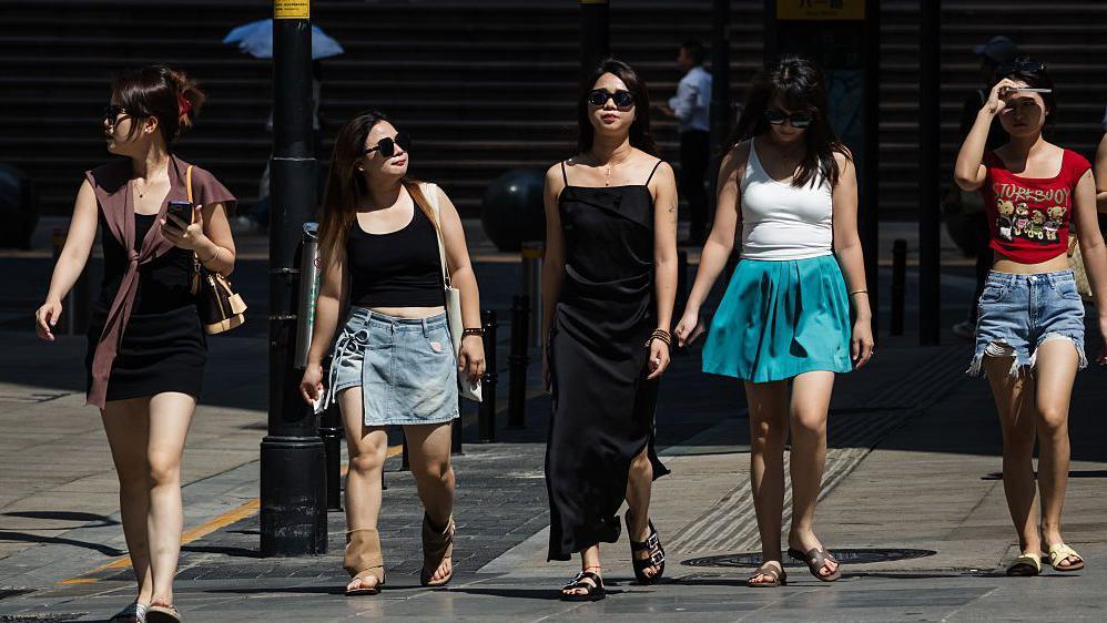 A group of young women wearing summer outfits walk through strong sunlight on a city street during a heatwave, as intense midday sun casts sharp shadows and reflects off the pavement, on June 16, 2025 in Chongqing, China.