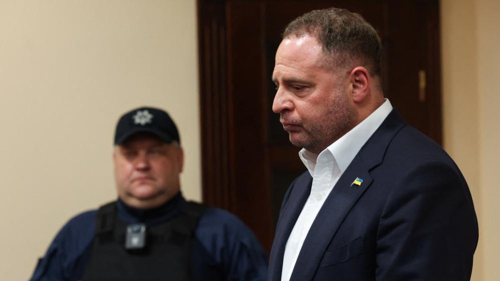 A man in a suit stands in court while a policeman looks on