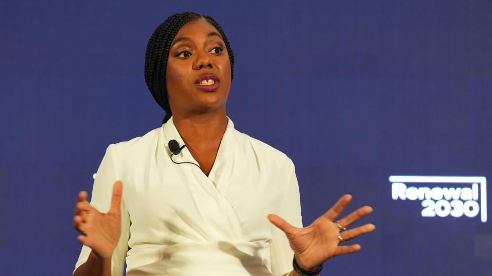 Kemi Badenoch speaks onstage at the launch of her leadership campaign. She is wearing a white blouse and holding her hands in front of her as she speaks. The stage background is blue. 